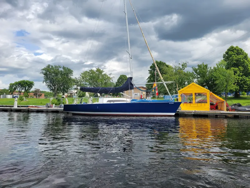 A navy blue racing sloop alongside the dock at Oromocto, a yellow tent and green trees behind, clouds overhead