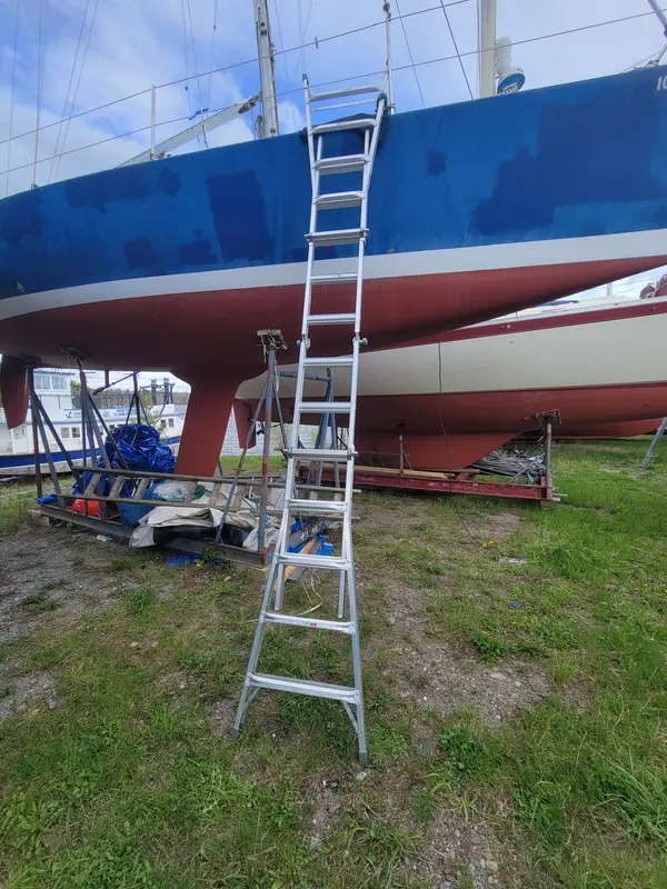 A blue hull with a red bottom on stands in a boatyard, a ladder against her side